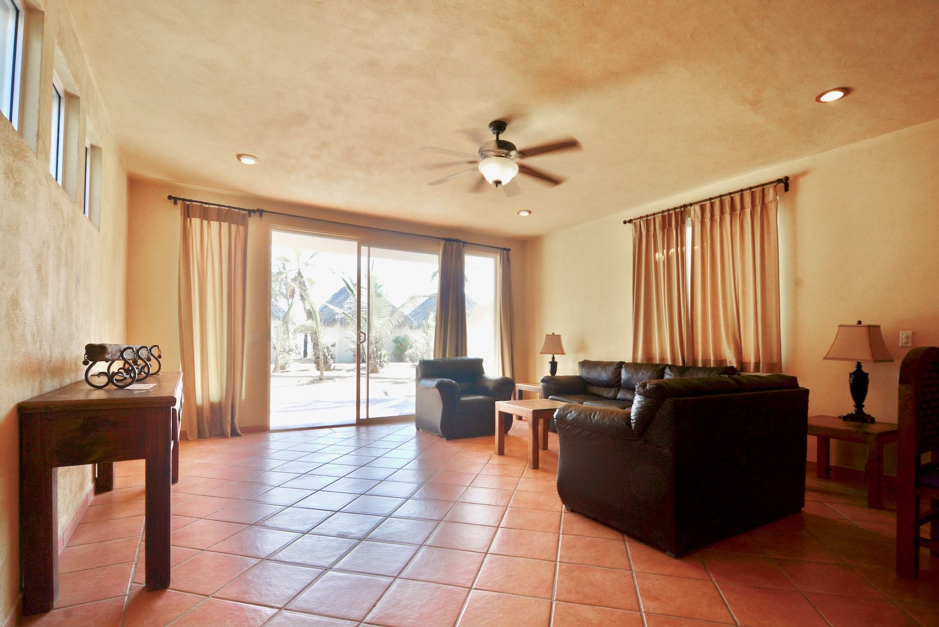 A spacious living room with a ceiling fan, a sofa, a coffee table, and a view of a tropical outdoor area through large sliding doors.