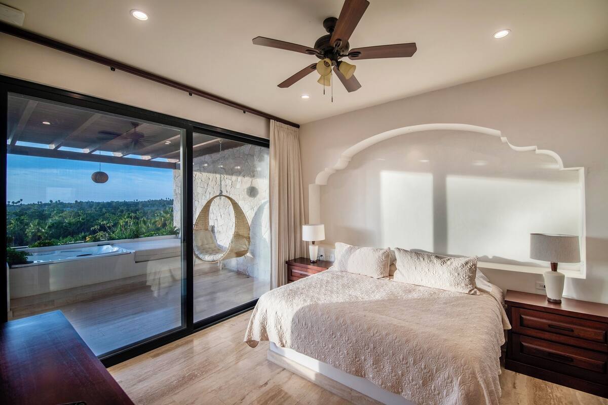 Een slaapkamer met een groot bed, een plafondventilator, een nachtkastje met lamp en uitzicht op een balkon met een hangstoel.