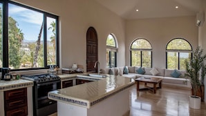 A spacious kitchen with a large island, a dining area with a sofa, and large windows offering a view of palm trees and hills. - Hacienda Todos Los Santos (Todos Santos)