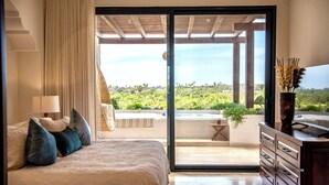 A bedroom with a bed, a dresser, a lamp, a curtain, and a view of a balcony with greenery. - Hacienda Todos Los Santos (Todos Santos)