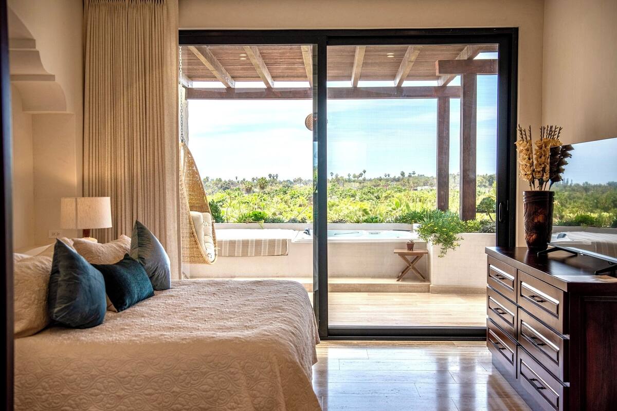 Een slaapkamer met een bed, een dressoir, een lamp, een gordijn en uitzicht op een balkon met groen.