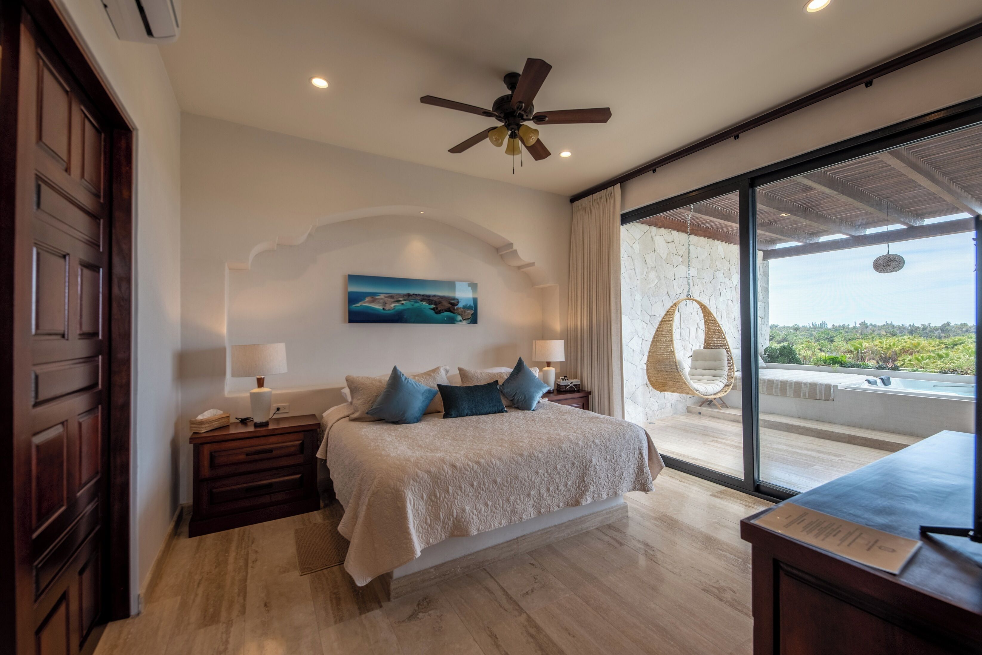 A bedroom with a bed, nightstands, a ceiling fan, a large window with a view, and a hanging chair.