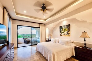 A bedroom with a large bed, a ceiling fan, a nightstand, a lamp, a TV, a dresser, and a sliding glass door leading to a balcony with a view.