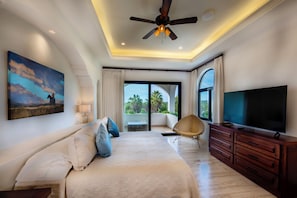 Un dormitorio con cama, televisor, ventilador de techo, ventana y un cuadro en la pared.