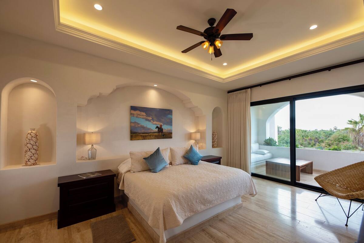Een slaapkamer met een bed, een plafondventilator, een nachtkastje, een lamp, een schilderij, een raam en uitzicht op de omgeving.