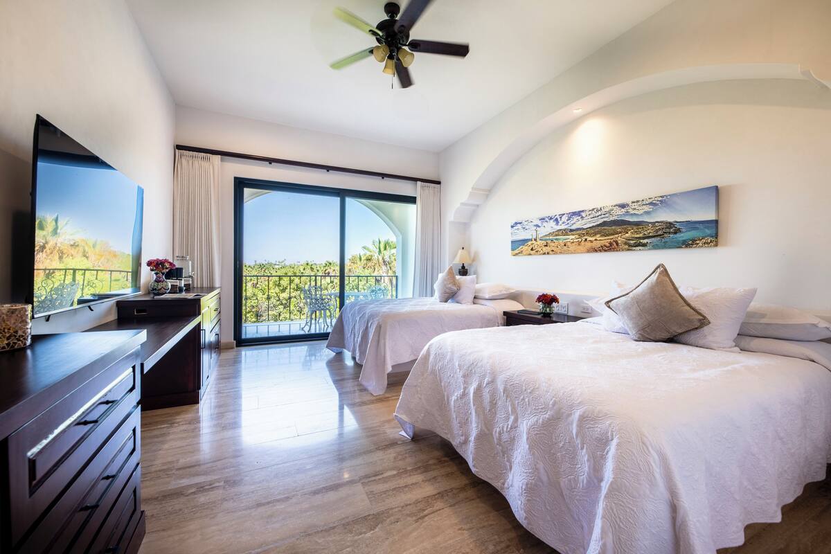 Een hotelkamer met twee bedden, een groot raam met uitzicht, een plafondventilator en een schilderij aan de muur.