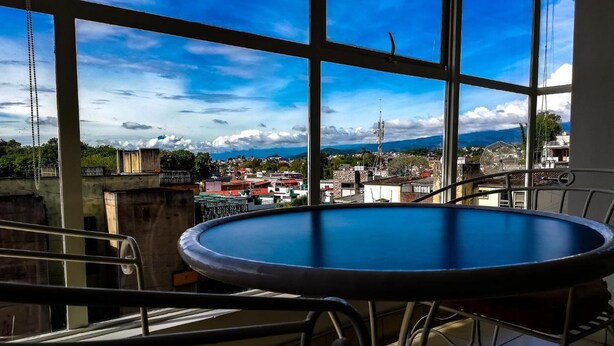 Un balcón con una mesa redonda y sillas, con vista a un paisaje urbano que incluye edificios y montañas.