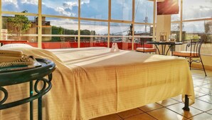 Una habitación con una cama, una mesa con sillas y ventanas amplias que ofrecen vistas de la ciudad.