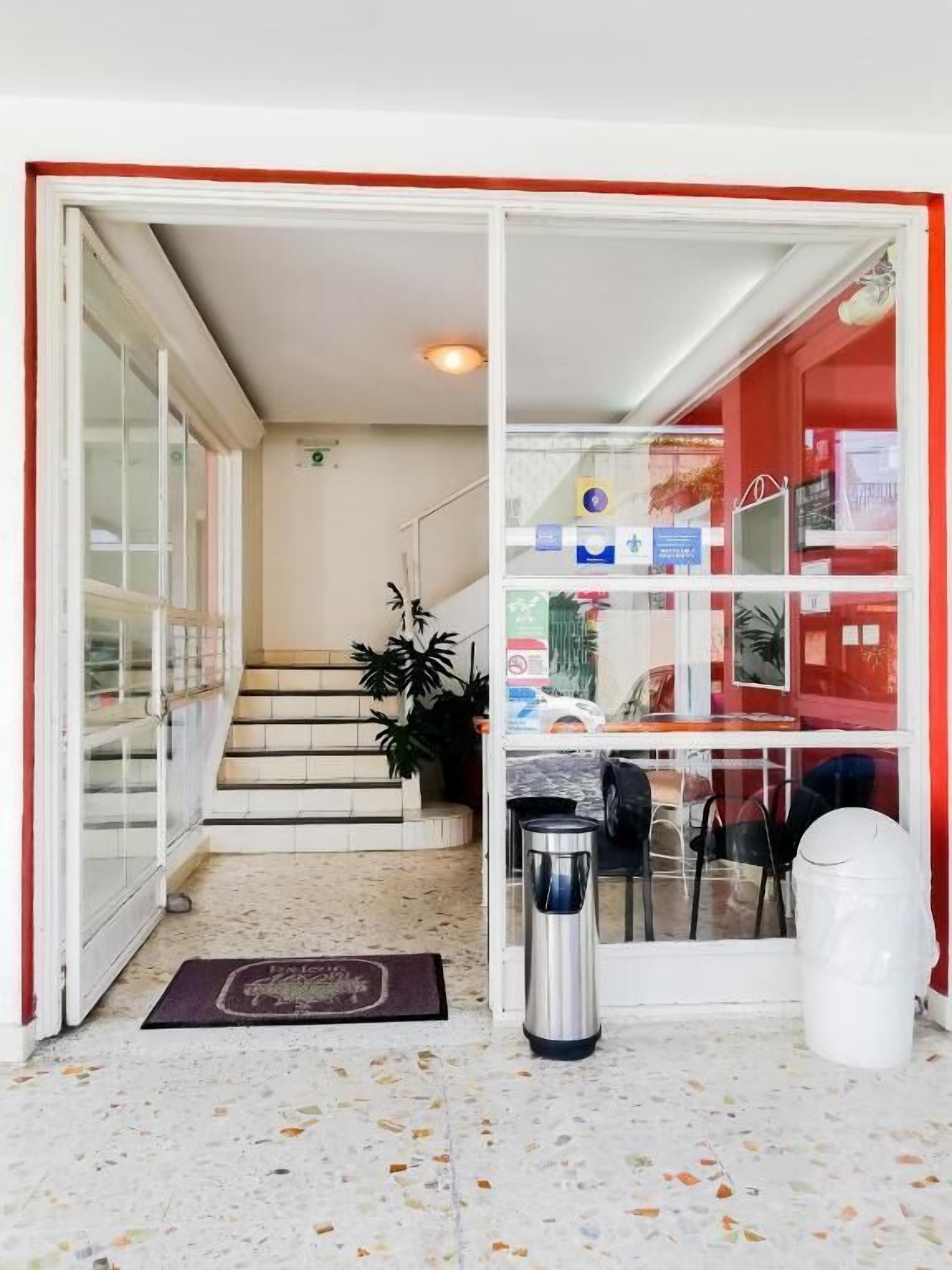 A hotel entrance with a glass door, a reception desk, a trash bin, and a staircase leading to the upper floor.