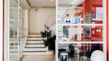 A hotel entrance with a glass door, a reception desk, a trash bin, and a staircase leading to the upper floor.