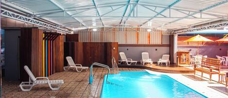 An indoor swimming pool with a tiled floor, lounge chairs, and a covered seating area.