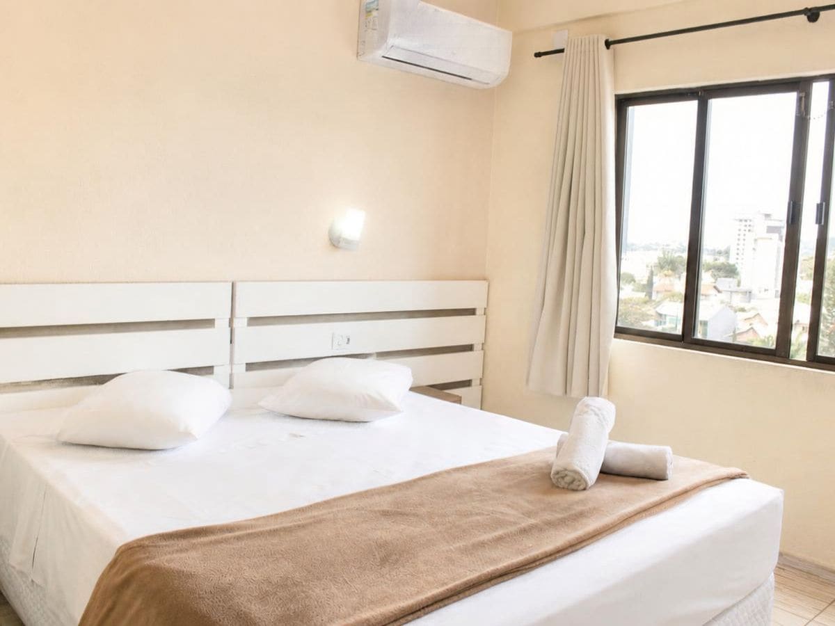 Una habitación de hotel con una cama, cortinas blancas, un ventanal con vistas y un equipo de aire acondicionado.