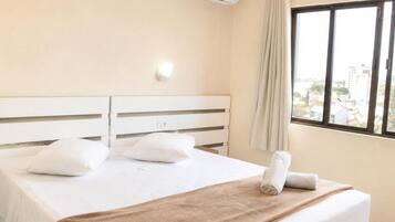 Una habitación de hotel con una cama, cortinas blancas, un ventanal con vistas y un equipo de aire acondicionado.
