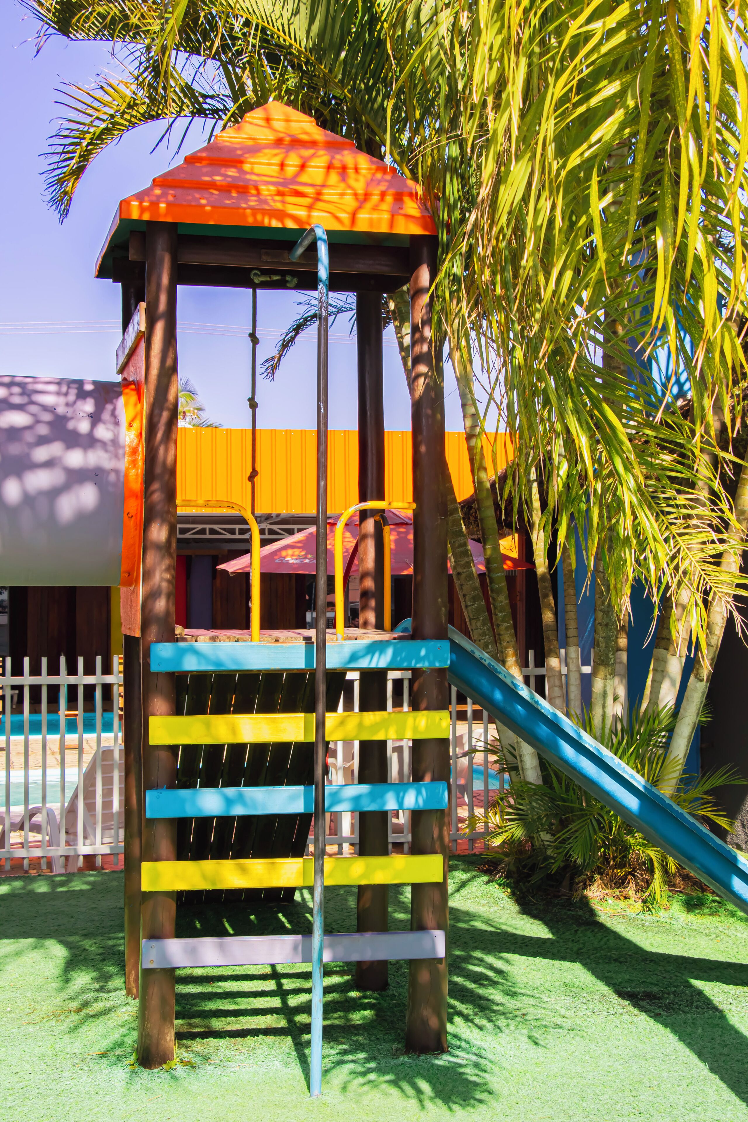 Une structure de jeux pour enfants comprenant un toboggan et des éléments d’escalade, entourée de palmiers et d’une clôture.