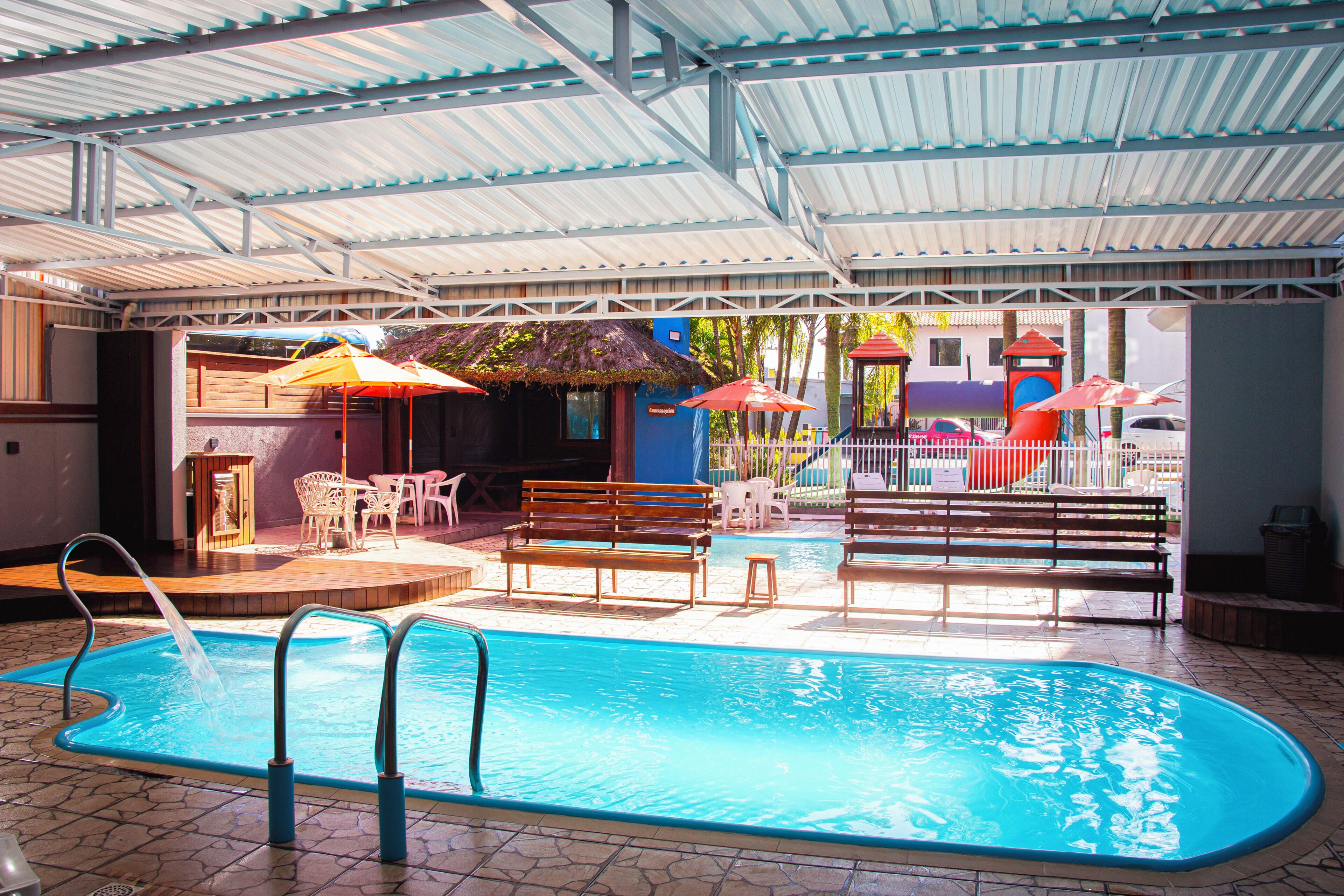 Piscina interior com chão em mosaico, rodeada por bancos e cadeiras de madeira, e uma área coberta com teto de metal.
