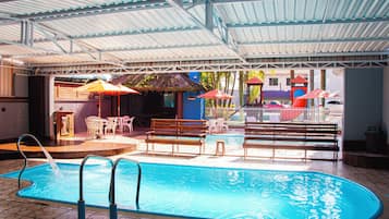 Piscina interior com chão em mosaico, rodeada por bancos e cadeiras de madeira, e uma área coberta com teto de metal.