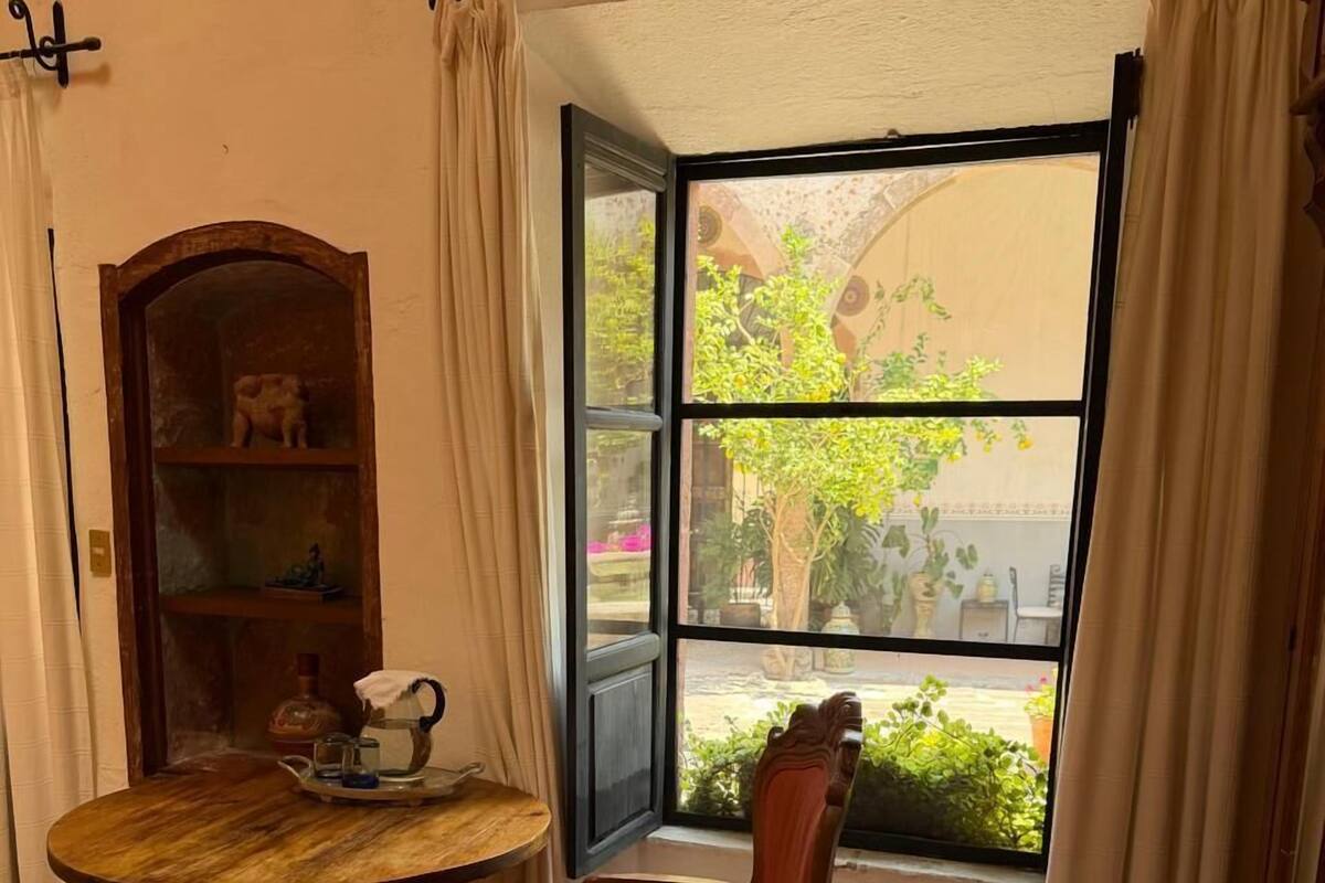 A room with a round wooden table, a red cushioned chair, a bookshelf, and a window with curtains.