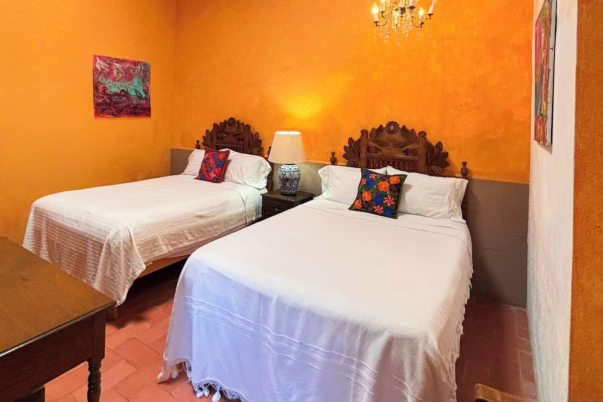 A room with two beds, a chandelier, a painting, and a wooden table.