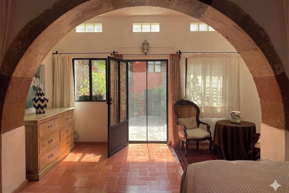 A room with a large arched doorway, a bed, a chair, a small table, and a dresser.