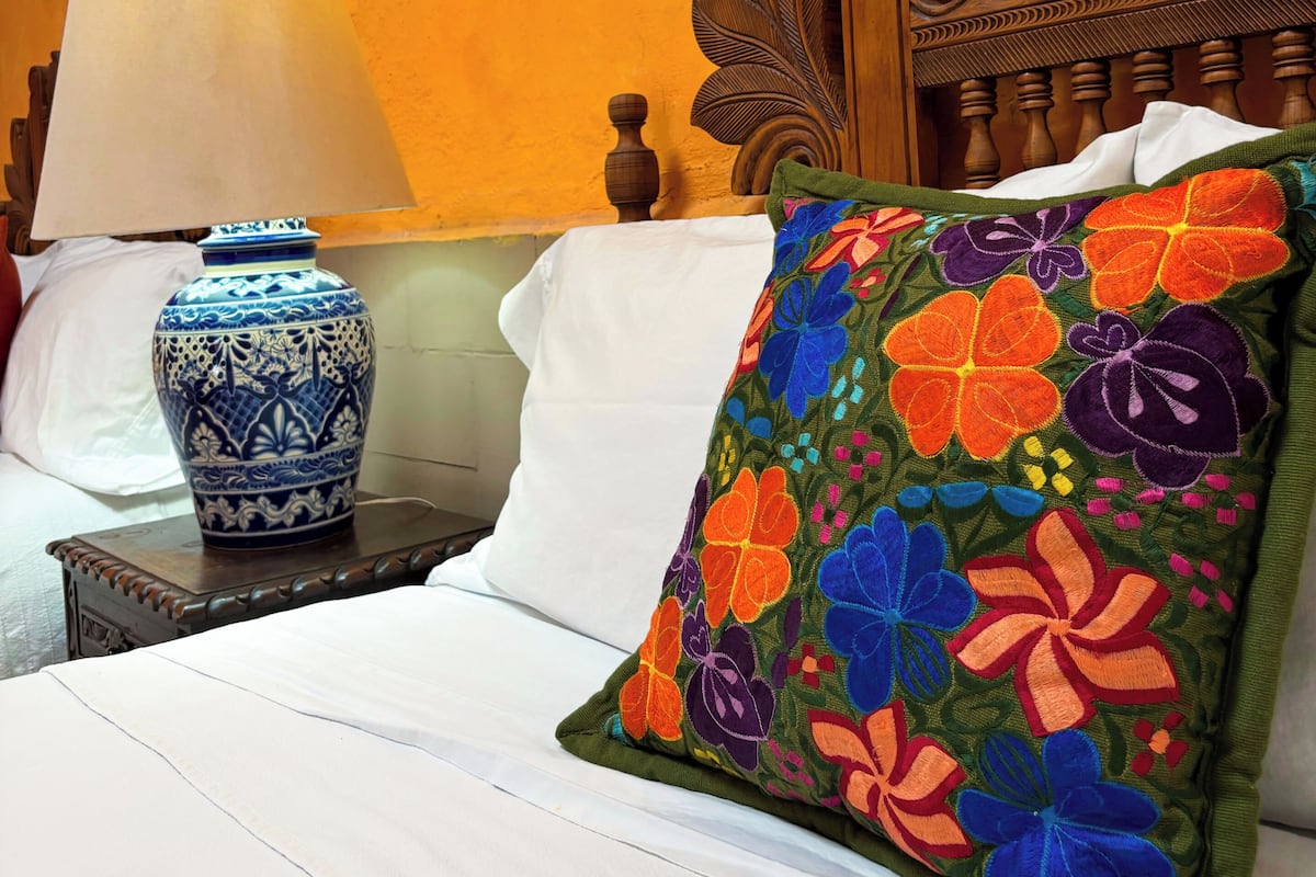 A neatly made bed with a colorful embroidered pillow, a blue and white vase, and a wooden headboard with a floral design.