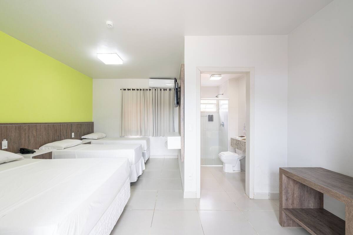 A hotel room with three beds, a wooden bench, a bathroom with a shower, and a toilet.