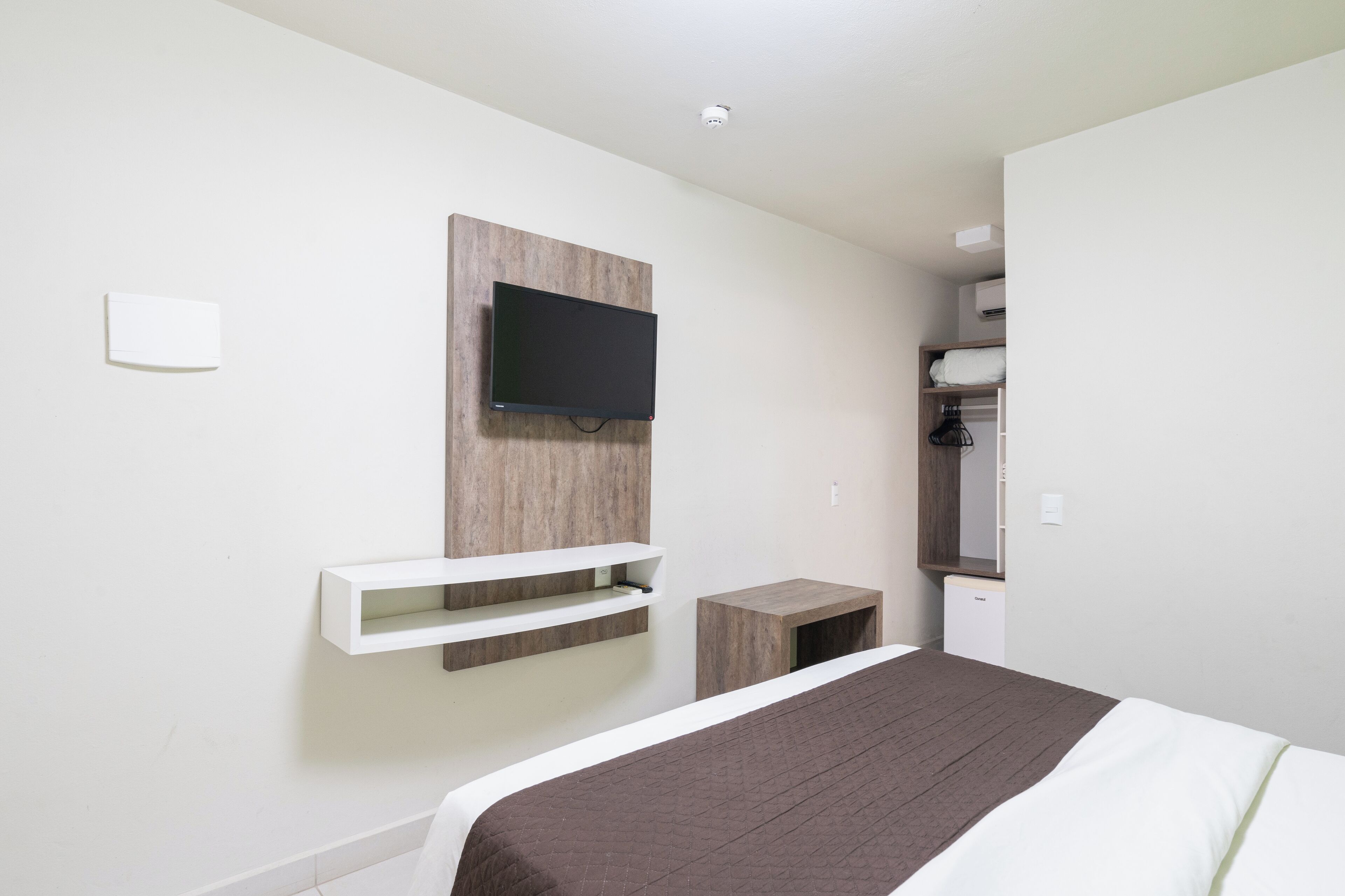 A modern hotel room with a bed, a mounted TV, a small bedside table, and a wardrobe.
