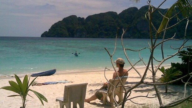 A beach with a chair and a small boat on the sand, a person sitting on the chair, and a view of the sea and a mountain in the background. - P.P. Blue Sky Resort (Ko Phi Phi)