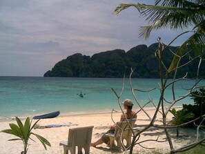 A beach with a chair and a small boat on the sand, a person sitting on the chair, and a view of the sea and a mountain in the background. - P.P. Blue Sky Resort (Ko Phi Phi)