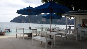 Un café junto a la playa con mesas blancas y sombrillas azules, con vistas a un mar tranquilo donde se ven barcos y acantilados rocosos al fondo.