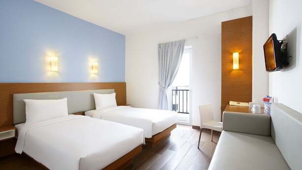 Ein Hotelzimmer mit zwei Betten, einem Schlafsofa, einem an der Wand montierten Fernseher und einem Fenster mit Vorhängen.