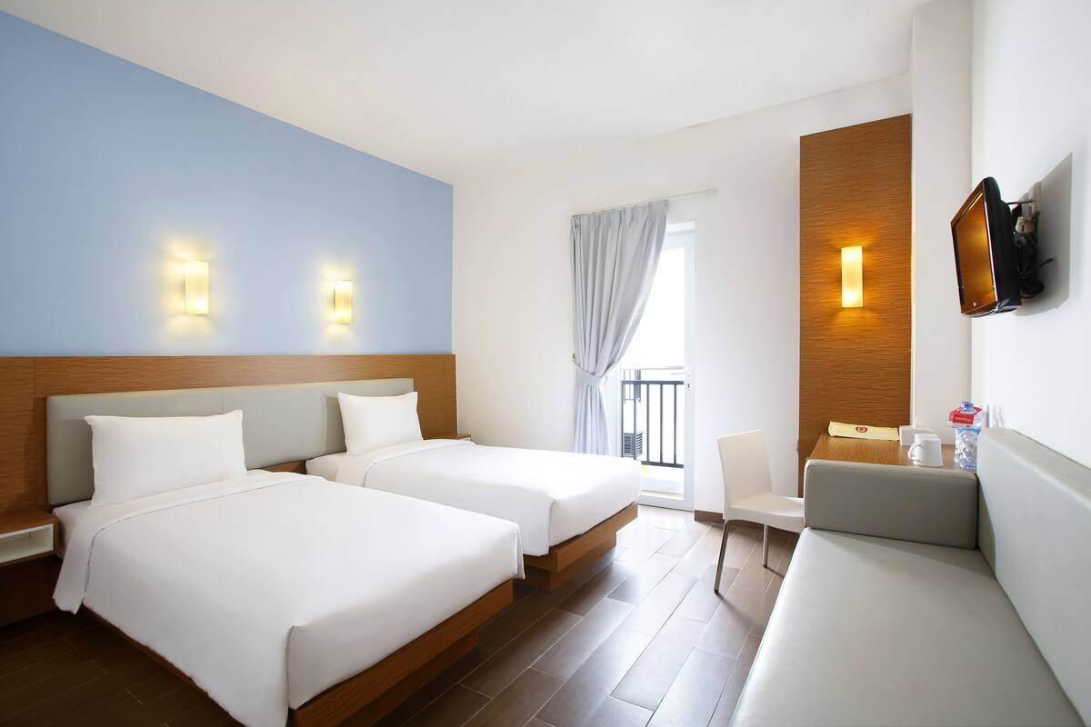 Habitación de hotel con dos camas, un sofá, un televisor montado en la pared y una ventana con cortinas.