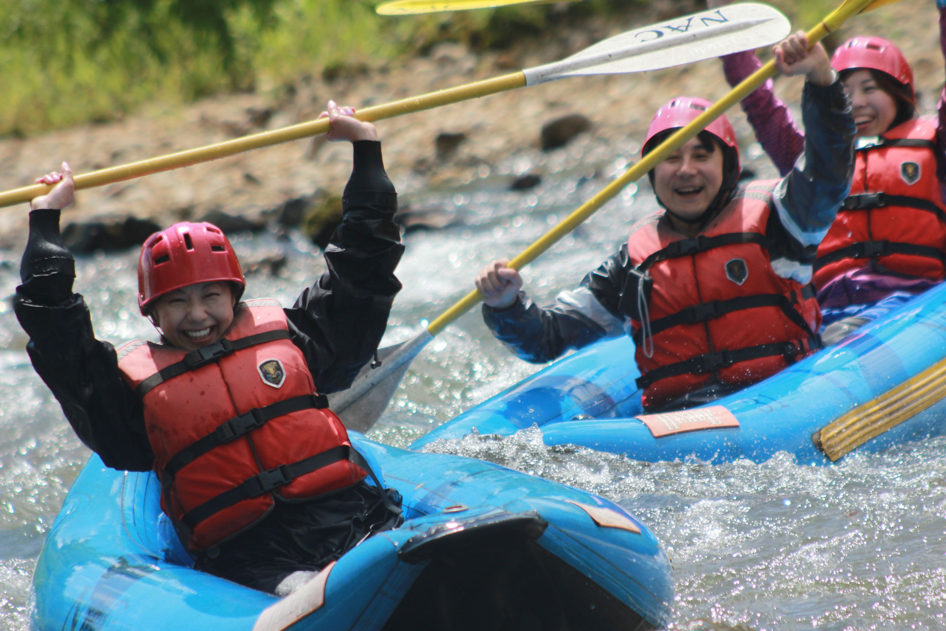 A group of people in a blue inflatable raft, each wearing a red life jacket and a helmet, paddling through a river.