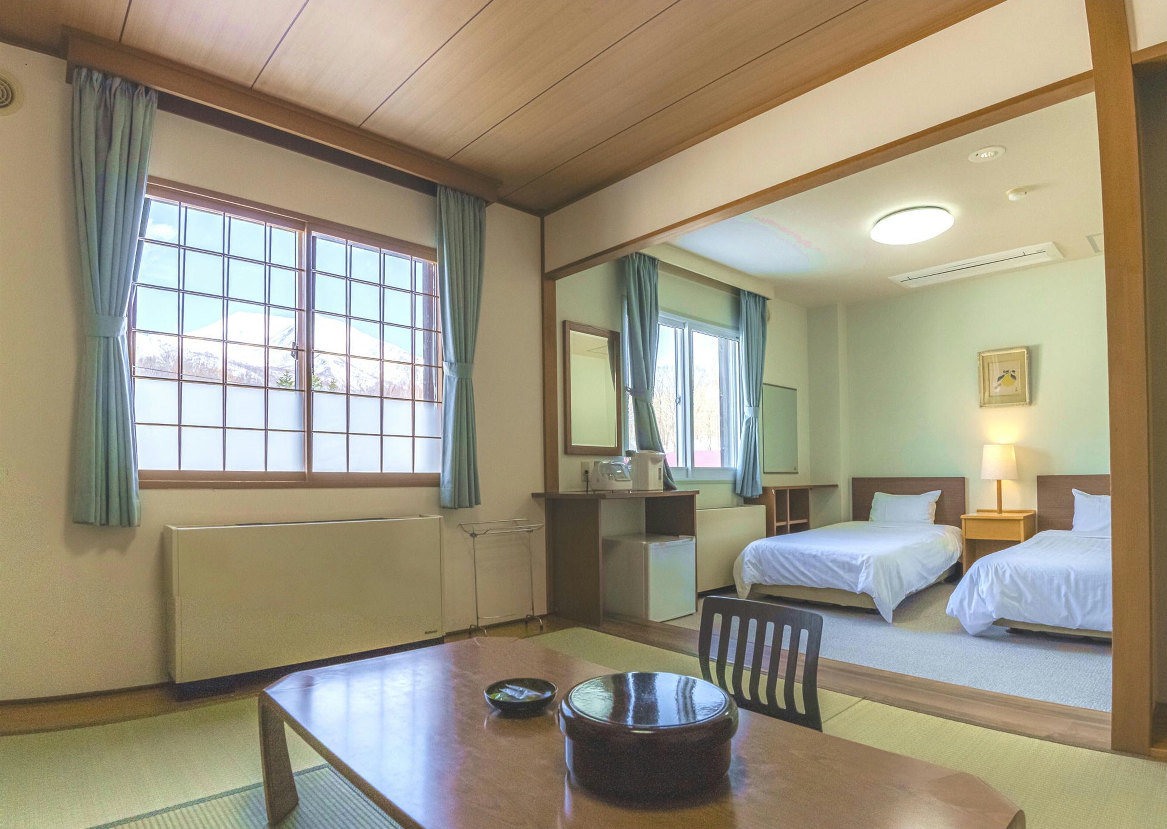 Una habitación de hotel con techo de madera, un ventanal con cortinas, un escritorio con un cuenco, dos camas, una mesita de noche con una lámpara y un espejo en la pared.