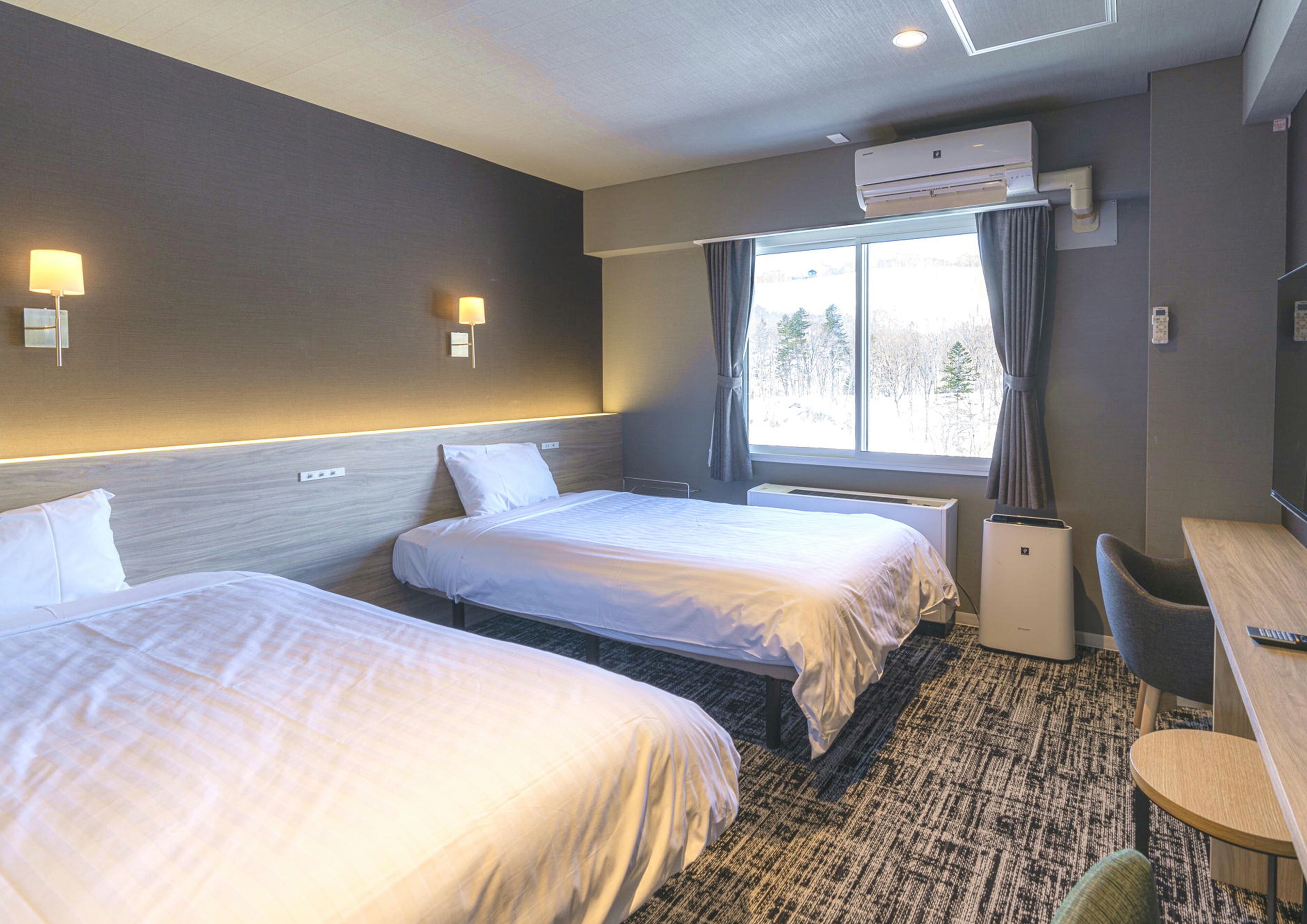 Ett hotellrum med två sängar, ett skrivbord, en stol, en luftkonditioneringsanläggning och ett fönster med gardiner.