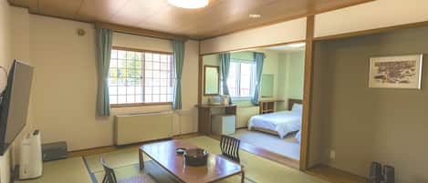 A traditional Japanese-style room with tatami flooring, a low table, a bed, a desk, and a window with curtains.