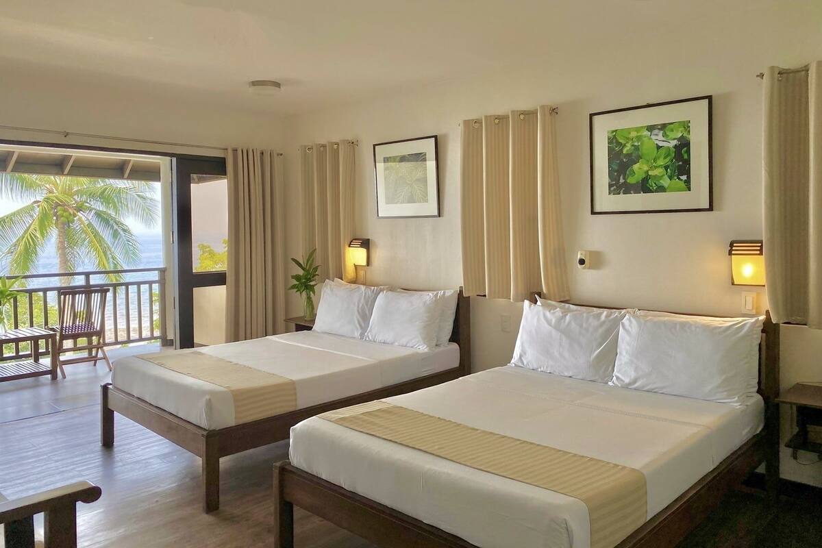 A hotel room with two beds, a balcony with a view of palm trees, and a small table.