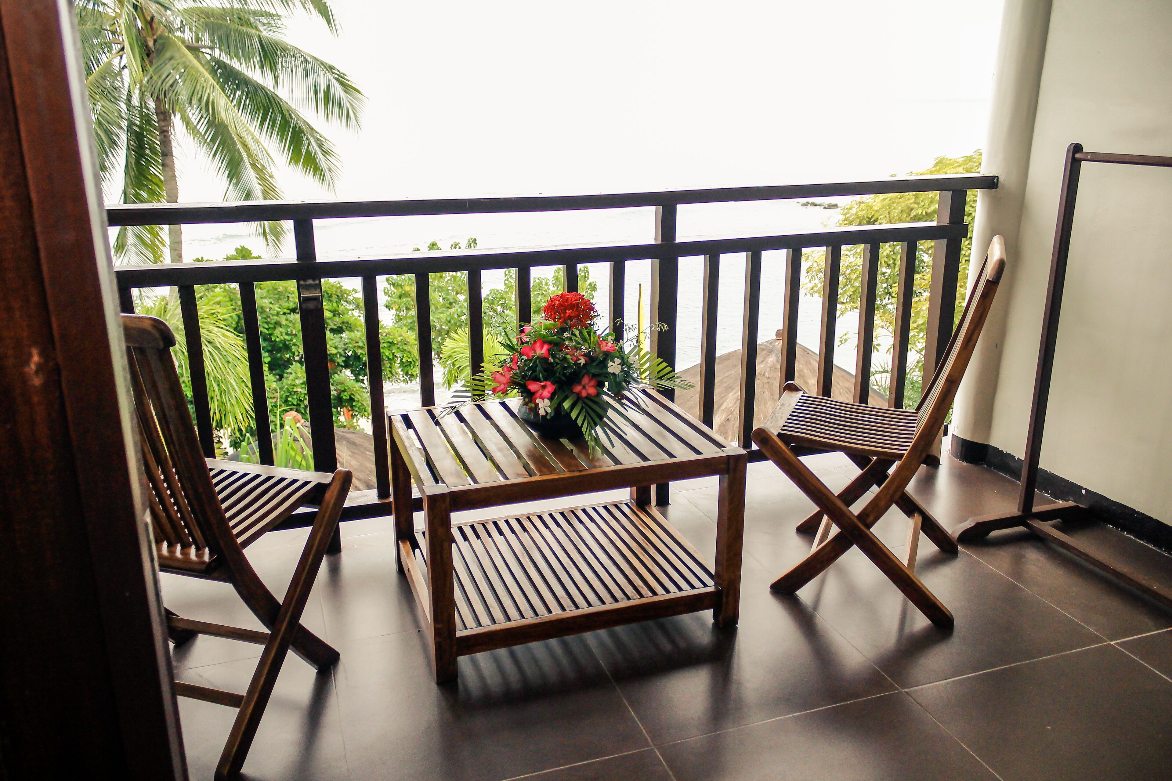 A balcony with two wooden chairs and a table, a flower vase, and a view of palm trees and greenery.