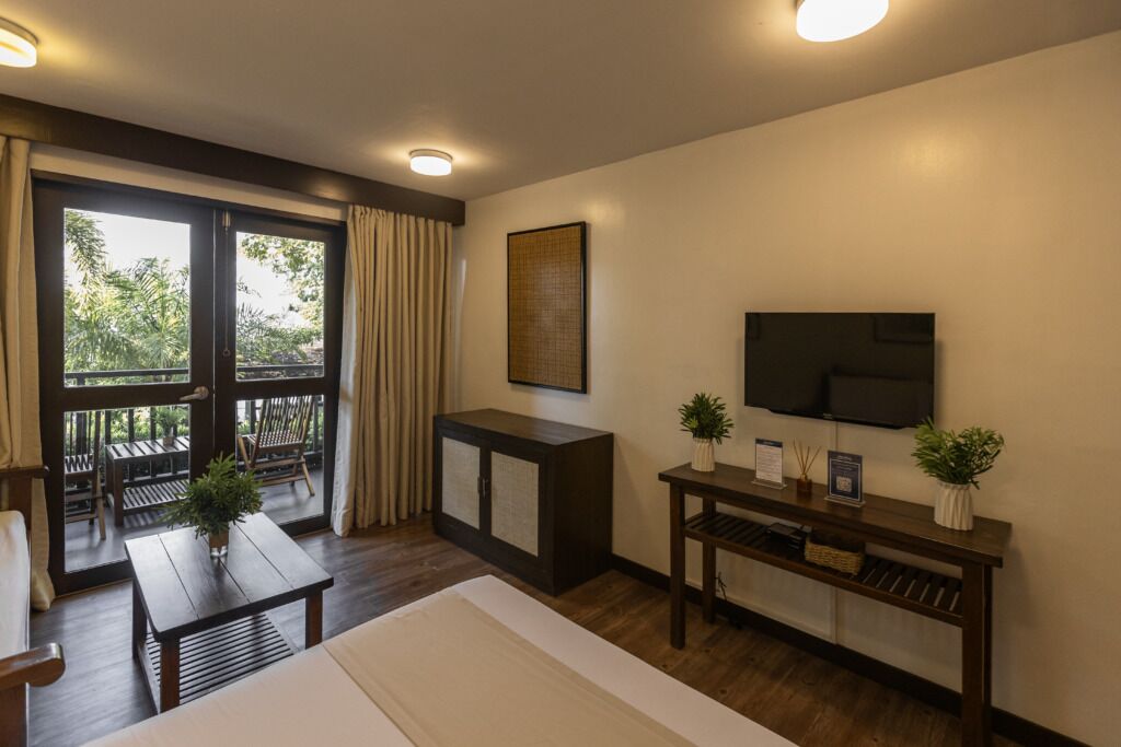 Una habitación con cama, escritorio, televisión y balcón con mobiliario exterior.