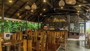 Un restaurante con mesas y sillas de madera, una pared de piedra con un letrero de neón y lámparas de mimbre colgantes.