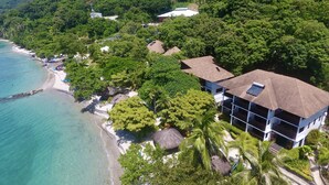 Vista aérea de una propiedad costera con playa, abundante vegetación y un edificio de techo inclinado.