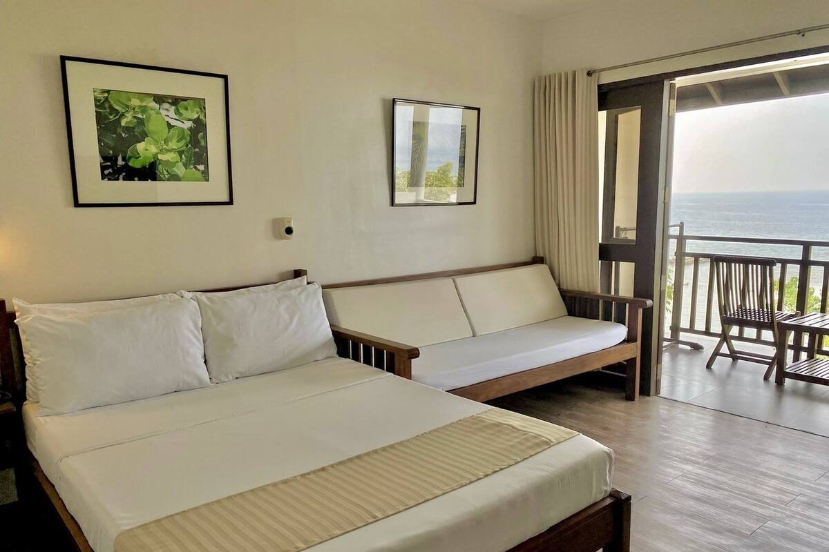 A hotel room with a bed, a sofa, a balcony with a view of the sea, and two framed pictures on the wall.