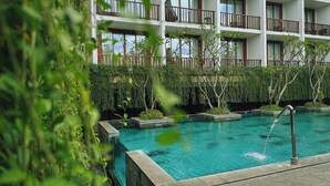 A pool area with a clear blue pool, surrounded by lush greenery and a multi-story building with balconies. - Grand Zuri Kuta Bali (Kuta)