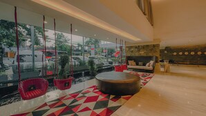 A modern lobby with a large window, a round wooden table, red wicker chairs, and a stone wall. - Grand Zuri Kuta Bali (Kuta)