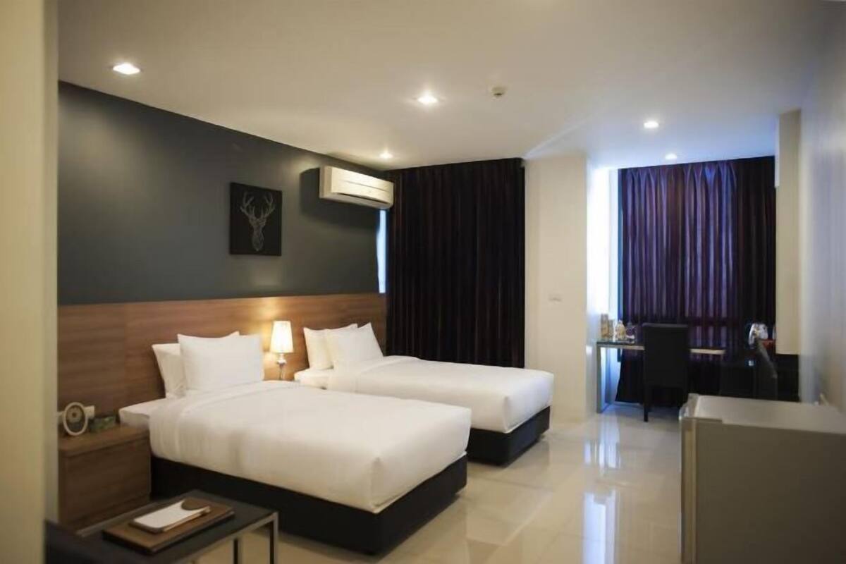 Habitación de hotel con dos camas, un escritorio, una silla, un equipo de aire acondicionado y una ventana con cortinas.