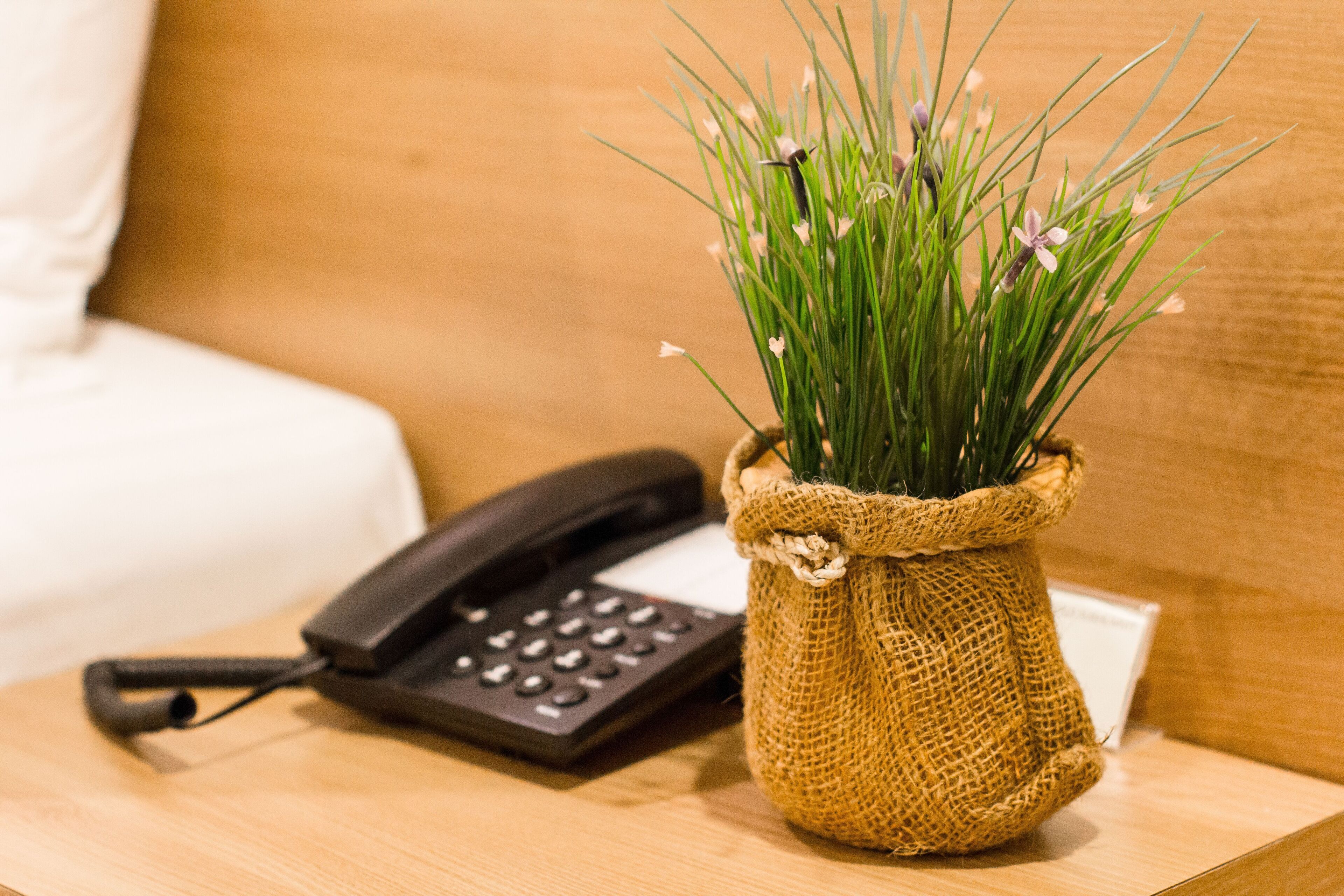 Ein kleiner Jutesack mit grünem Gras und Blumen auf einer Holztischplatte neben einem schwarzen Telefon.