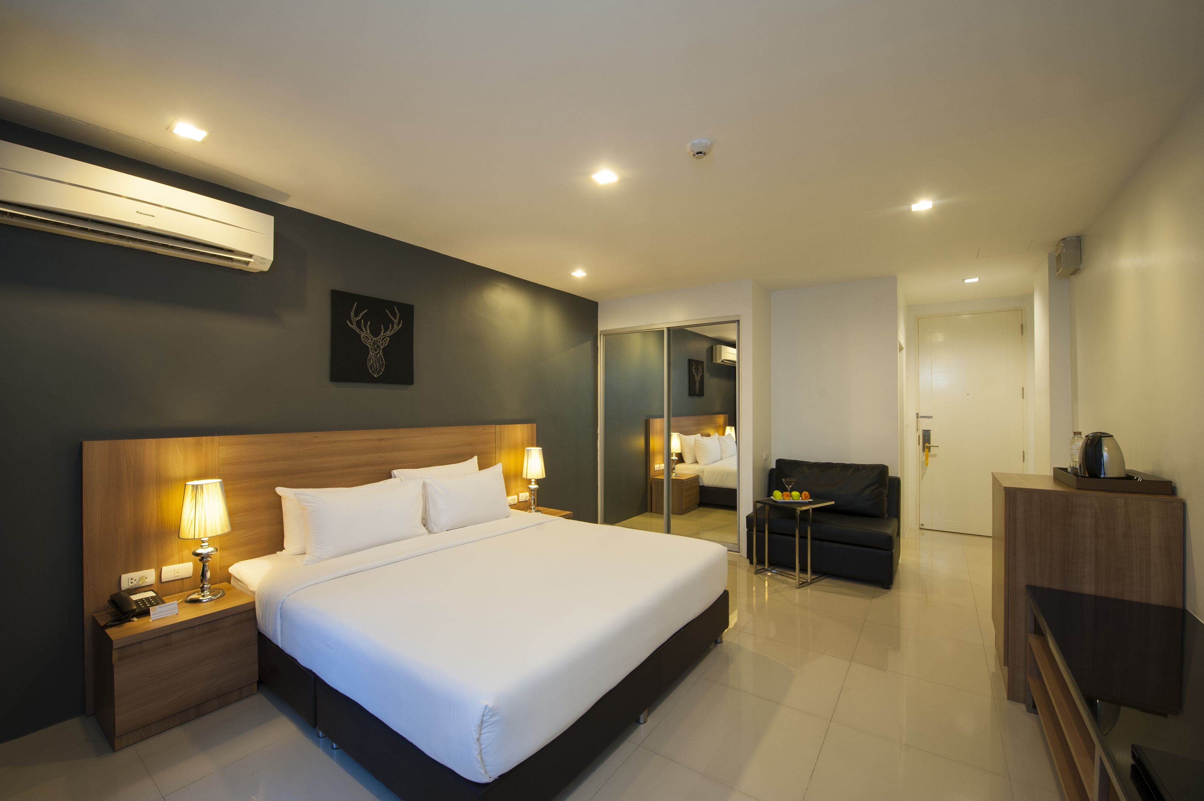 A modern hotel room with a large bed, bedside lamps, a wooden headboard, a small table, a sofa, and a TV.