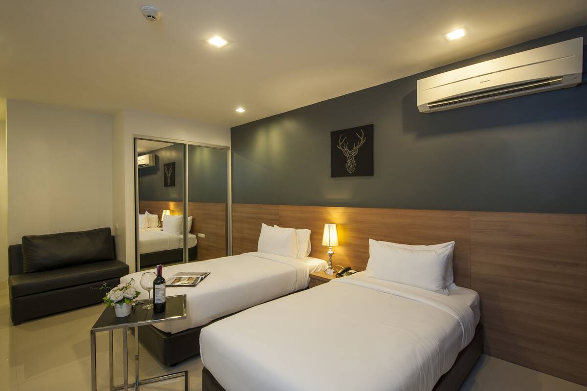 Habitación de hotel con dos camas, un sofá, una mesita con una botella de vino y un aire acondicionado en la pared.