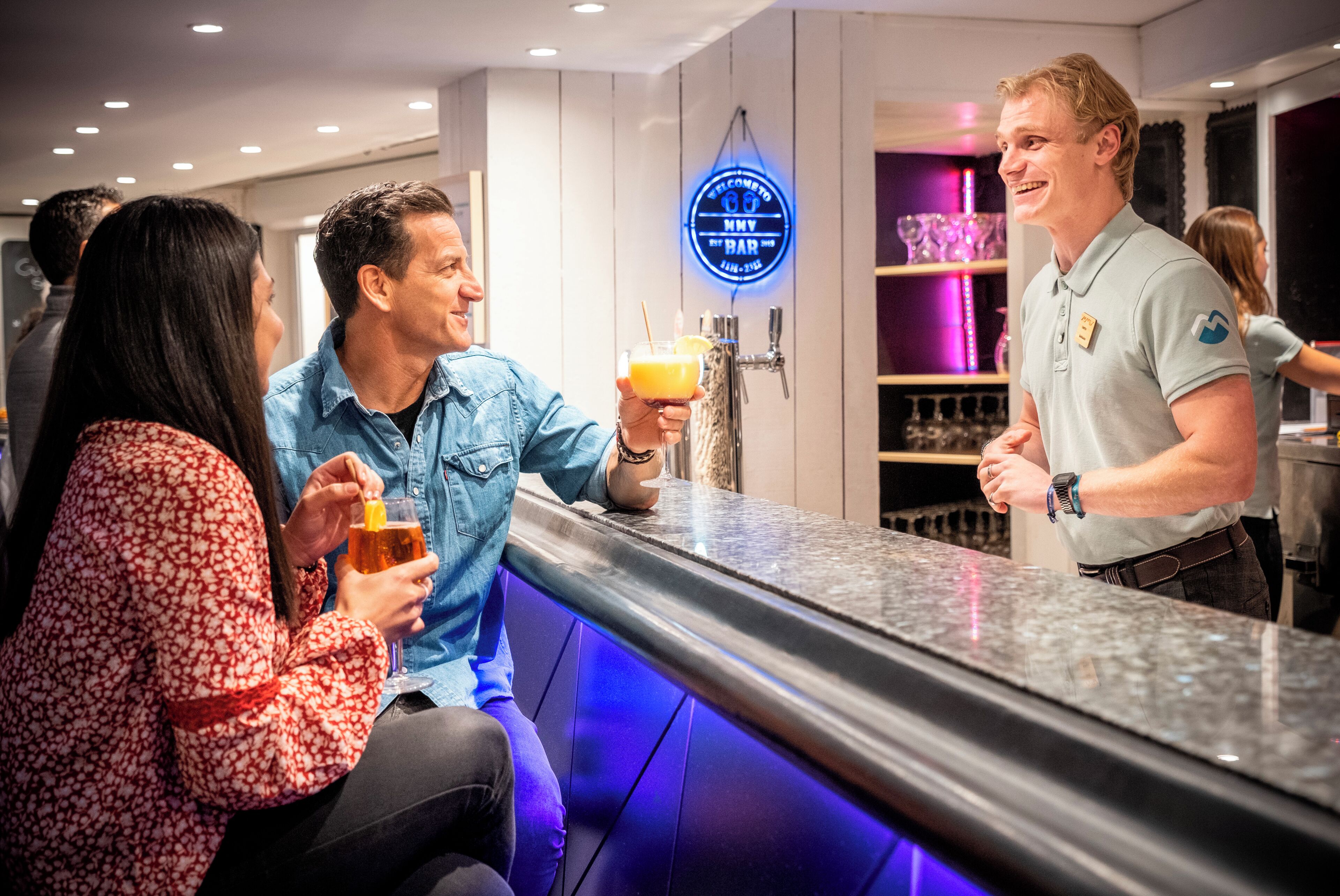A bar area with a neon sign, a counter, and people seated.