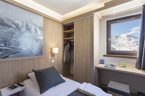 Quarto de hotel com cama, mesa de cabeceira, uma grande janela com vista para montanhas, guarda-roupa e um quadro de uma paisagem nevada.