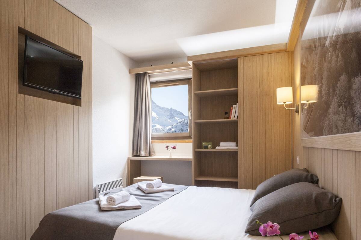 Habitación de hotel con cama, televisor montado en la pared, ventana con vista a la montaña y repisa con libros y un jarrón de flores.